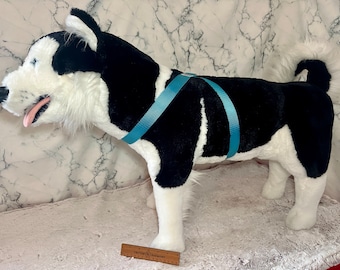 May include: A black and white plush husky dog with a blue harness. The dog is standing on a white fluffy surface.
