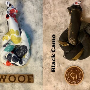 May include: Two dog toys, one with a paw print pattern and the other with a black camo pattern. Both toys are knotted and have a brown leather tag with the text "Critter Harmony" on it. The paw print toy has the word "WOOF" written on a wooden sign.