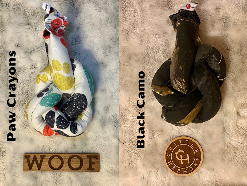 May include: Two dog toys, one with a paw print pattern and the other with a black camo pattern. Both toys are knotted and have a brown leather tag with the text "Critter Harmony" on it. The paw print toy has the word "WOOF" written on a wooden sign.