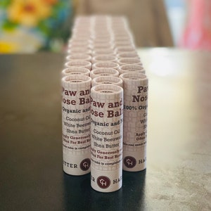 Op de afbeelding: Een verzameling cilindrische tubes met het opschrift "Paw and Nose Balm". De tubes bevatten ingrediënten zoals kokosolie en sheaboter. De tubes zijn in nette rijen op een donkere ondergrond gerangschikt.