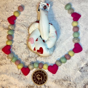 May include: A white knotted fabric toy with pink and red hearts printed on it. The toy is surrounded by a garland of pink, green, and yellow felt balls. The garland has a wooden tag with the text "Critter Harmony" on it.