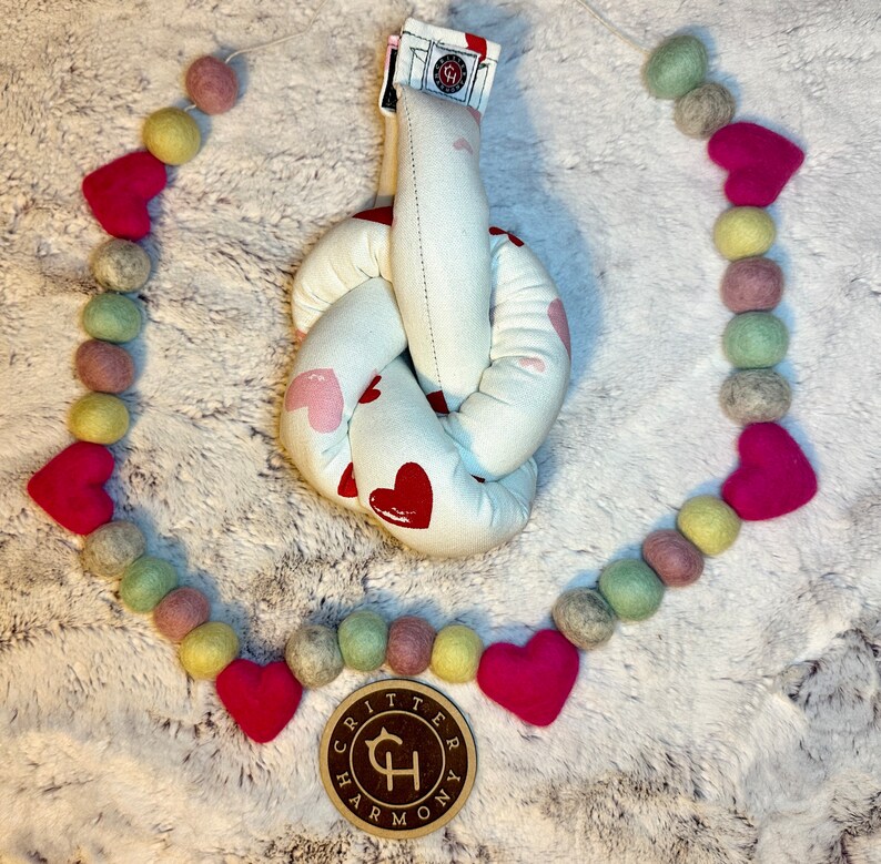 May include: A white knotted fabric toy with pink and red hearts printed on it. The toy is surrounded by a garland of pink, green, and yellow felt balls. The garland has a wooden tag with the text "Critter Harmony" on it.