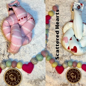 May include: Two knotted fabric heart-shaped toys. The toy on the left is pink with white stripes and small red hearts. The toy on the right is white with scattered pink and red hearts. Both toys have a brown tag with the text "Critter Harmony" and a stylized "CH" logo.