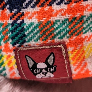 May include: A red, white, and blue plaid fabric with a small patch featuring a black and white bulldog with the text "CH CH" on its collar.