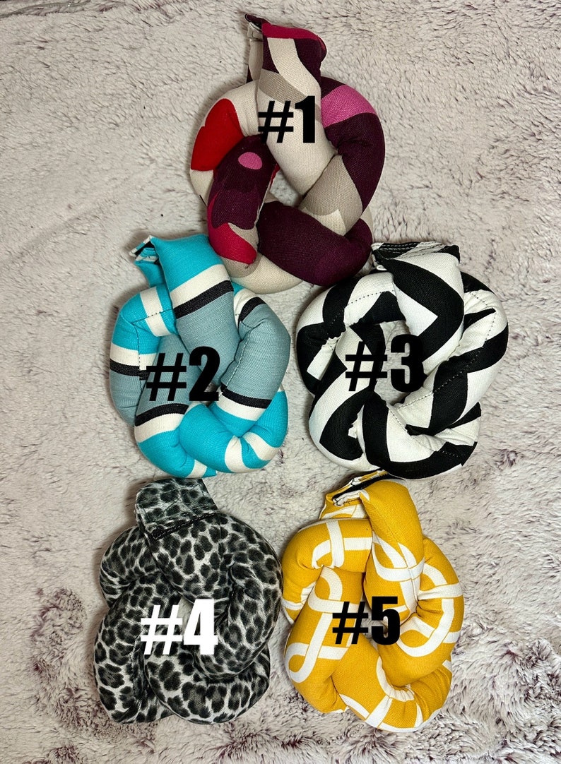 May include: Five knotted fabric pillows in different patterns. The pillows are numbered 1 through 5. Pillow #1 is pink, white, and brown with a red heart. Pillow #2 is turquoise, white, and gray. Pillow #3 is black and white. Pillow #4 is gray and black with a leopard print. Pillow #5 is yellow with a white pattern.