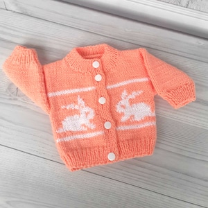 May include: A peach-colored knitted cardigan with white bunny designs and white buttons.