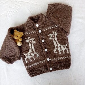 May include: A brown knitted baby cardigan with white giraffe designs and white buttons. The cardigan is lying on a white surface with a small brown teddy bear.