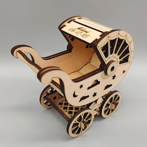 May include: A wooden baby carriage with intricate cut-out designs. The carriage features decorative wheels, stars, and clouds. The date "06-17-25" is engraved on the top. The carriage is made of light-colored wood.