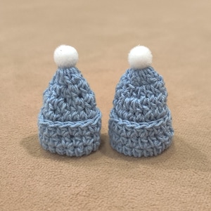 May include: Two small blue crocheted hats with white pom-poms. The hats are made with a textured stitch and have a rounded shape.