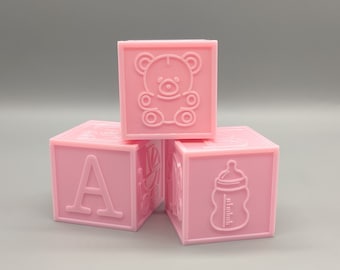 May include: Three pink baby blocks with different designs. One block has the letter A, another has a teddy bear, and the third has a baby bottle.