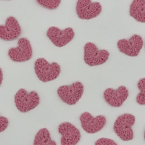 May include: A collection of small, individual heart-shaped motifs crafted from light pink yarn using a crochet technique. Each heart features a textured, looped stitch pattern and is arranged in a scattered, non-uniform pattern across the frame.