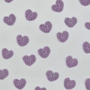 May include: A collection of small, light purple crocheted hearts arranged in a scattered pattern. Each heart features a textured, yarn-based construction with a visible stitch pattern, creating a uniform appearance across all individual pieces.