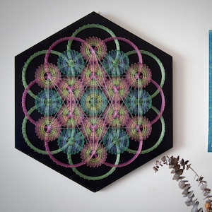 May include: A hexagonal wooden frame with a colorful string art design. The design features a geometric pattern with circles and triangles in shades of pink, green, and blue. The background is black.