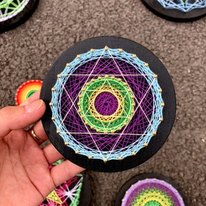 May include: A circular string art piece with a black background. The string art features a multi-colored design with concentric circles and a star shape in the center. The colors used are purple, blue, green, yellow, and gold.