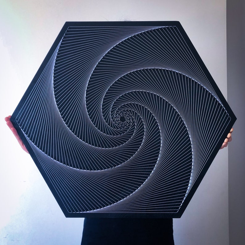May include: A black hexagonal wall art piece with a white string art design of a spiral. The spiral is made of many thin lines that create a hypnotic effect.
