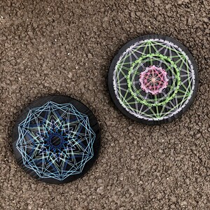 K&ouml;nnte beinhalten: Zwei schwarze, runde Untersetzer aus Holz mit geometrischen String-Art-Designs. Einer hat blaue und wei&szlig;e F&auml;den, der andere gr&uuml;ne, rosa und wei&szlig;e. Die Untersetzer liegen auf einer strukturierten, dunkelgrauen Oberfl&auml;che.