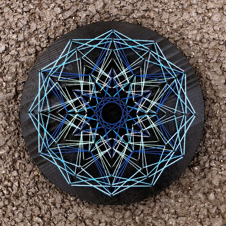 K&ouml;nnte beinhalten: Ein rundes, schwarzes Holz-String-Art-St&uuml;ck mit einem geometrischen Design. Das Kunstwerk verwendet hellblaue, wei&szlig;e und dunkelblaue F&auml;den, um ein symmetrisches Muster zu erzeugen. Das St&uuml;ck wird auf einer strukturierten Oberfl&auml;che pr&auml;sentiert.