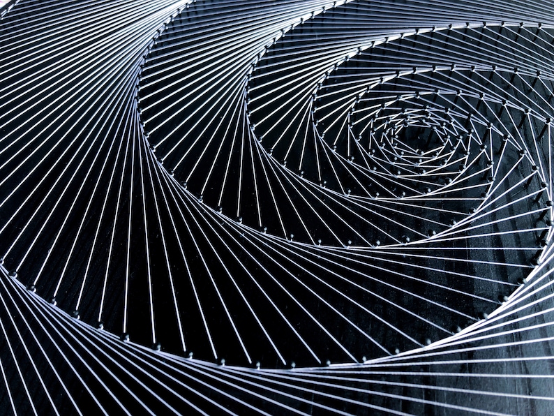 May include: A close-up of a string art piece with a spiral design. The string is white and the background is black. The string is wrapped around nails that are hammered into the black surface.