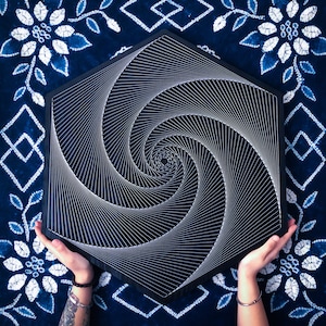Könnte beinhalten: Ein schwarzes sechseckiges Holzbrett mit einem weißen String-Art-Design einer Spirale. Das Brett wird von zwei Händen gehalten.