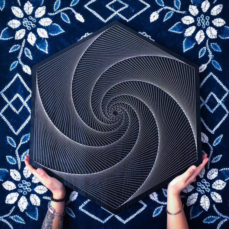 May include: A black hexagonal wooden board with a white string art design of a spiral. The board is held by two hands.