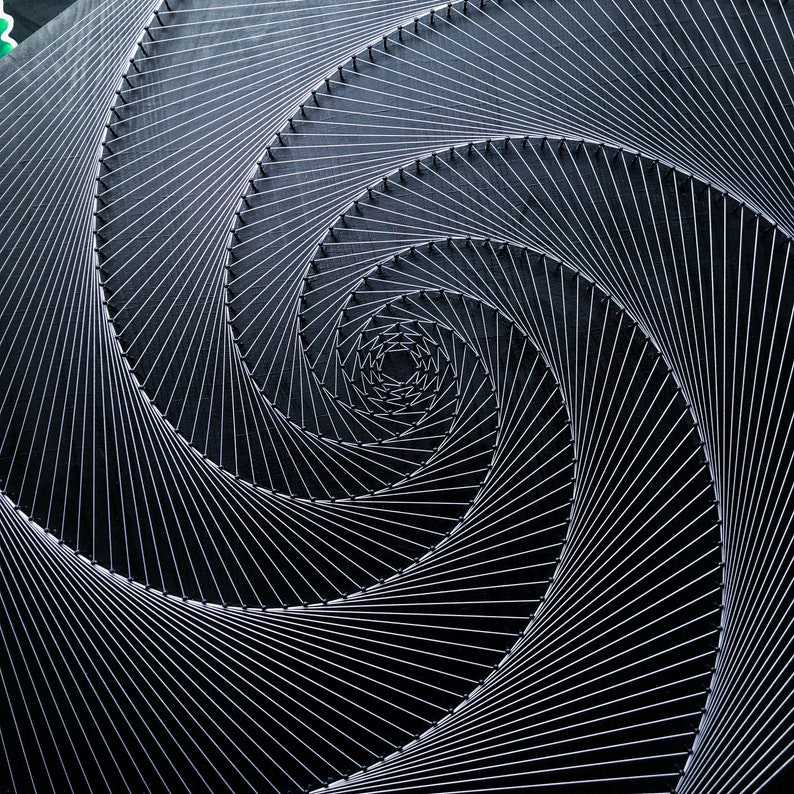 May include: A black and white abstract art piece made with string and nails. The string is arranged in a spiral pattern, creating a sense of depth and movement.