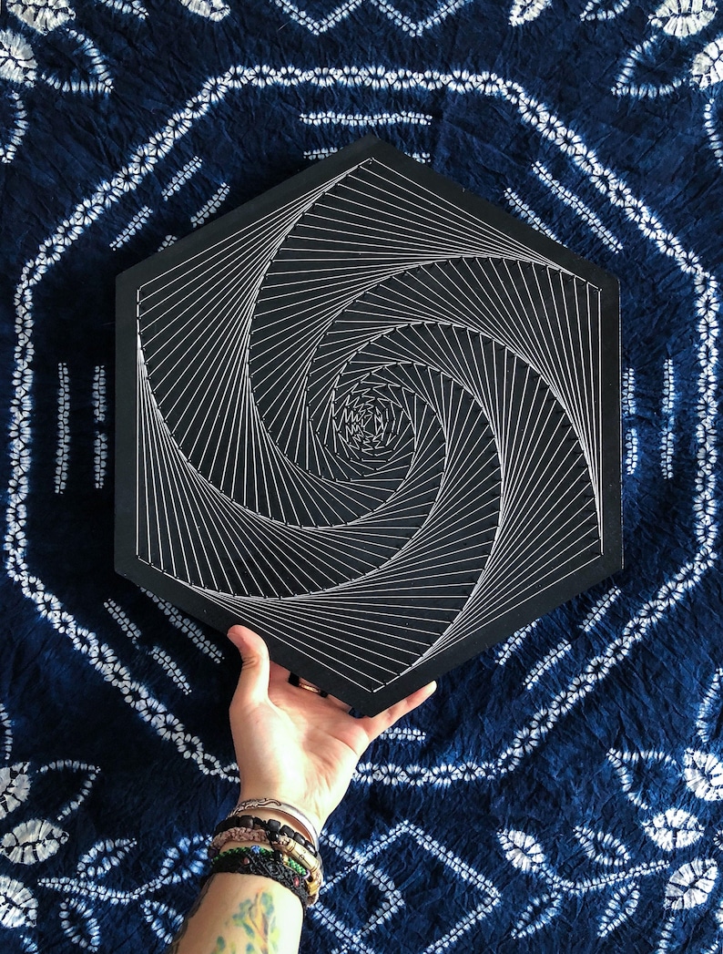 May include: A black hexagonal wooden board with a white string art design of a spiral. The spiral is made of many lines that converge in the center of the board.