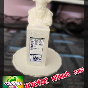 Peut inclure: Un étui blanc imprimé en 3D avec un buste de Viking sur le dessus. L'étui a un écran affichant des graphiques et du texte, et repose sur une base ronde. Le texte "BJORN ultimate case" est affiché en rouge.