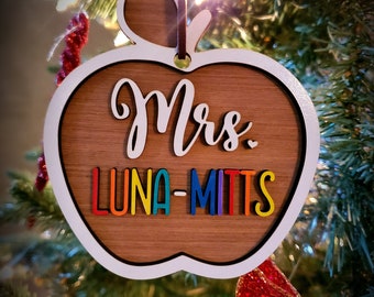 Può includere: Addobbo natalizio in legno a forma di mela con il testo "Mrs. Luna-Mitts" in colori arcobaleno.