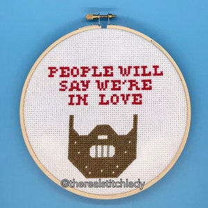 May include: A white embroidery hoop with the text "PEOPLE WILL SAY WE'RE IN LOVE" in red and a brown stitched image of a mask with a metal grate. The hoop is hanging on a blue wall. The text "@thereal stitchlady" is at the bottom of the hoop.