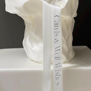May include: A white satin fabric container with a draped, gathered appearance. A long, smooth white satin ribbon hangs vertically down the front, featuring the black printed text "Cards & Well Wishes" in a clean, serif font style.