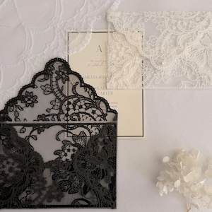 Puede incluir: Un sobre de encaje negro con un intrincado patrón floral y un sobre de encaje blanco a juego con un pequeño cierre de perla. Una tarjeta de invitación color crema con texto negro es parcialmente visible tras el sobre de encaje negro.