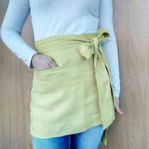 Könnte beinhalten: Hellgelbe Schürze mit Tasche und langen Bindebändern, seitlich zu einer Schleife gebunden. Die Person trägt ein langärmliges weißes Oberteil und blaue Jeans. Die Schürze ist aus weichem Stoff.