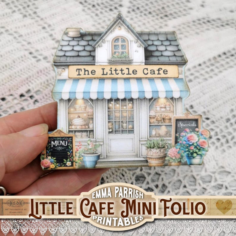 May include: A miniature cafe model with a blue and white striped awning, labeled "The Little Cafe." The facade includes a door, windows, and a chalkboard sign that says "Menu." The model is decorated with flowers and potted plants.