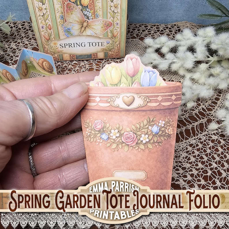 May include: A decorative paper flower pot with a floral garland and tulips. The pot is peach-colored with a heart design. The text "Spring Garden Tote Journal Folio Printables" is at the bottom.