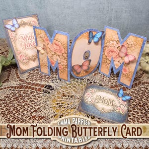 Puede incluir: Un juego de tarjetas decorativas "MOM" con detalles florales y mariposas. Las letras son de color marrón claro con un borde azul y presentan un patrón de rosas. El juego incluye una tarjeta con las palabras "With Love" y una tarjeta pequeña con la palabra "Mom".
