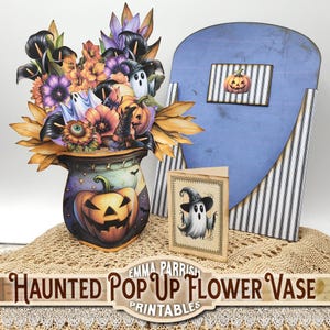 May include: A Halloween pop-up flower vase featuring a pumpkin design, filled with paper flowers, ghosts, and pumpkins. A blue backdrop with a pumpkin and a card with a ghost illustration complete the scene. The text reads "Haunted Pop Up Flower Vase Printables".