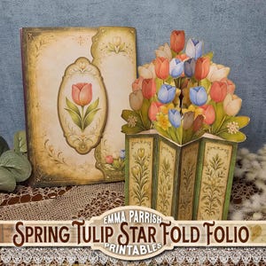 May include: A decorative paper craft featuring a pop-up tulip arrangement and a matching book-style folio. The tulips are in shades of red, orange, and blue. The folio and the base of the pop-up have floral designs and the text "Spring Tulip Star Fold Folio Printables".