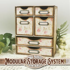 May include: A white, six-drawer storage unit with a floral pattern and gold accents. The drawers have labels with gold trim. The unit is labeled "Emma Parrish Modular Storage System Printables".
