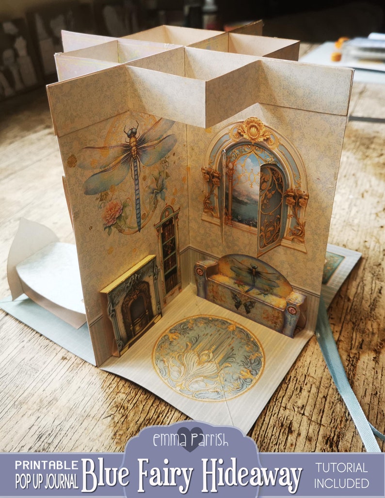 Puede incluir: Un diario emergente imprimible titulado "Blue Fairy Hideaway" con un tutorial incluido. El diario presenta un escondite de hadas azul y dorado con una chimenea, un banco y una lib&eacute;lula.