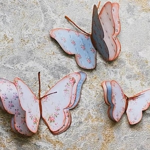 May include: Three paper butterflies with floral patterns in shades of blue, pink, and white. The butterflies are layered and have a 3D effect.