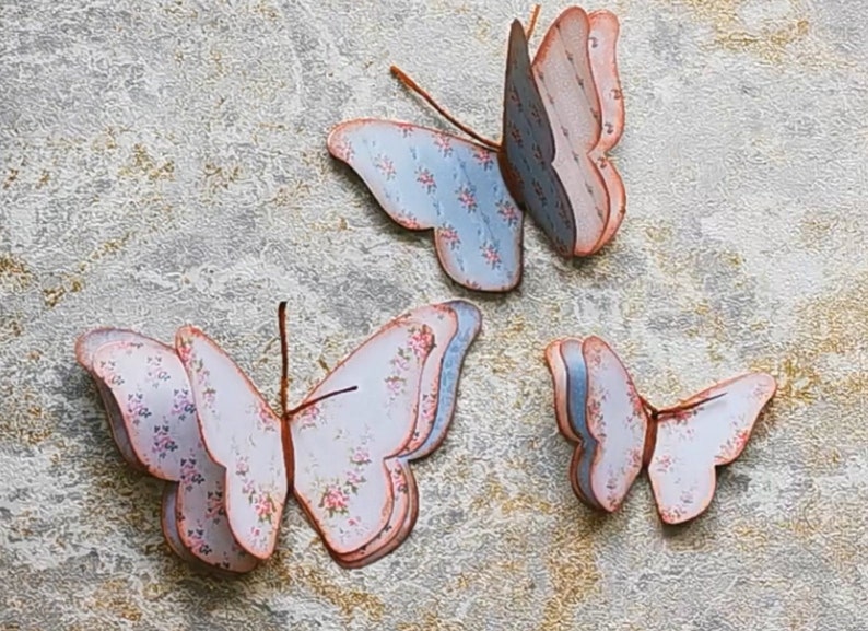 May include: Three paper butterflies with floral patterns in shades of blue, pink, and white. The butterflies are layered and have a 3D effect.