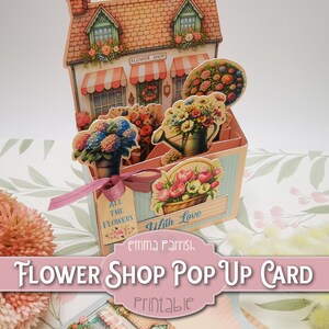Może przedstawiać: Drukowana rozkładana kartka z motywem kwiaciarni z czerwonym dachem, białym daszkiem i szyldem z napisem "Flower Shop". Kartka jest ozdobiona kwiatami i konewką. Na kartce znajduje się r&oacute;wnież tekst "With Love" i "Emma Parrish".