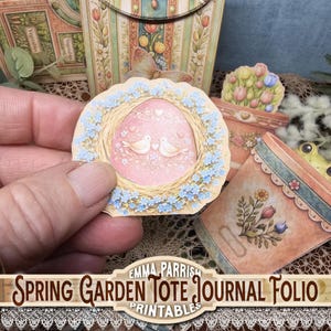 May include: A paper craft featuring a pink egg-shaped design with two white doves, surrounded by a wreath of blue flowers. The image is part of a spring garden tote journal folio, with additional floral and garden-themed elements.