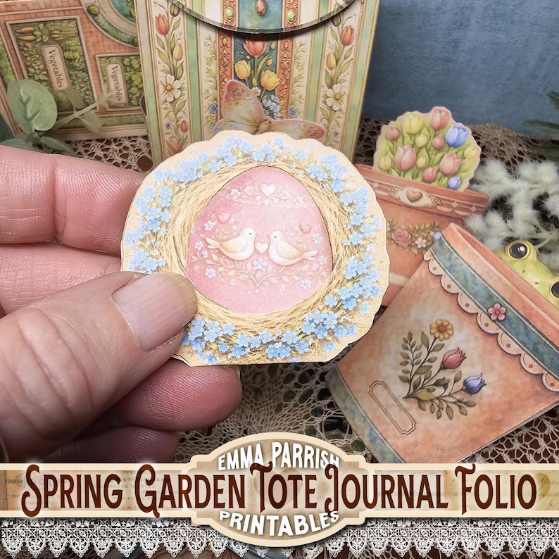 May include: A paper craft featuring a pink egg-shaped design with two white doves, surrounded by a wreath of blue flowers. The image is part of a spring garden tote journal folio, with additional floral and garden-themed elements.