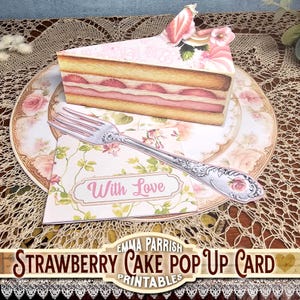 May include: A strawberry cake pop-up card with a slice of cake, a fork, and a card that says "With Love." The cake has pink frosting and strawberry slices. The card is on a floral plate.