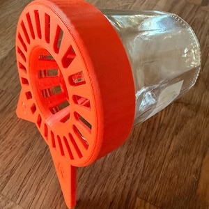 Op de afbeelding: Een oranje 3D-geprinte pottenhouder met een cirkelvormig ontwerp en een heldere glazen pot. De houder heeft een reeks verticale sleuven en een basis voor stabiliteit. De pot rust op een houten oppervlak.