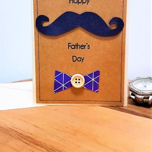 May include: A Father's Day card with a navy blue mustache and bow tie. The card has the words "Happy Father's Day" printed on it. The bow tie is purple with white geometric lines and a wooden button.