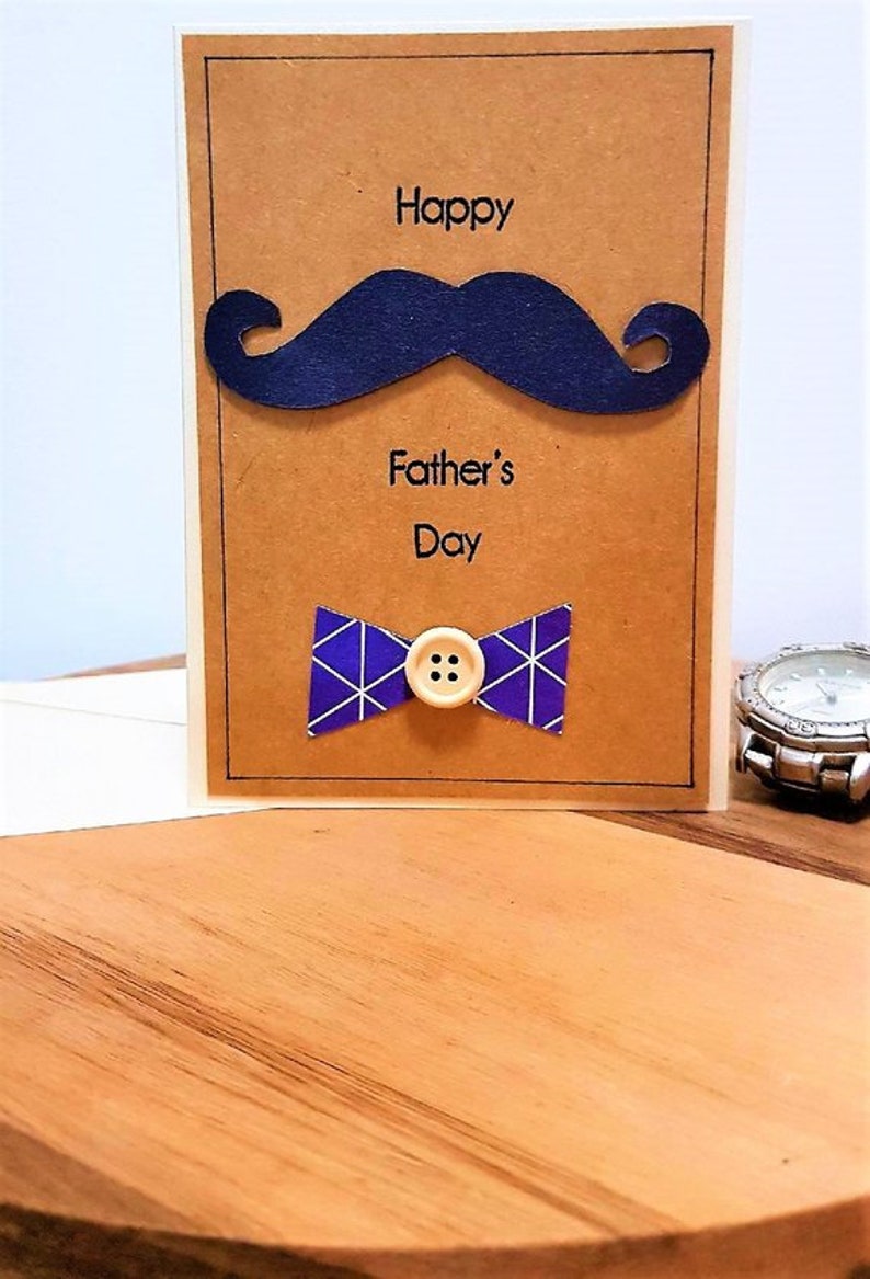 May include: A Father's Day card with a navy blue mustache and bow tie. The card has the words "Happy Father's Day" printed on it. The bow tie is purple with white geometric lines and a wooden button.
