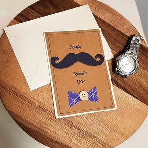 May include: A Father's Day card with a navy blue mustache and bow tie design, with the words "Happy Father's Day". The card is on a wooden surface, next to a white envelope and a silver wristwatch.