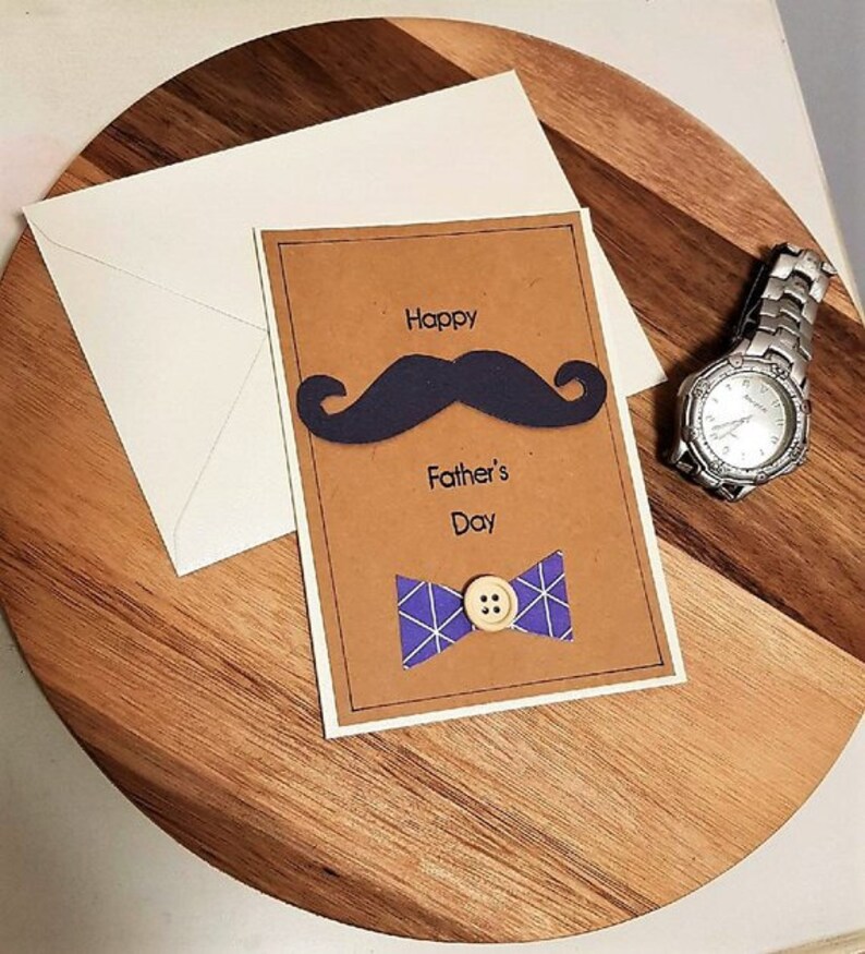 May include: A Father's Day card with a navy blue mustache and bow tie design, with the words "Happy Father's Day". The card is on a wooden surface, next to a white envelope and a silver wristwatch.